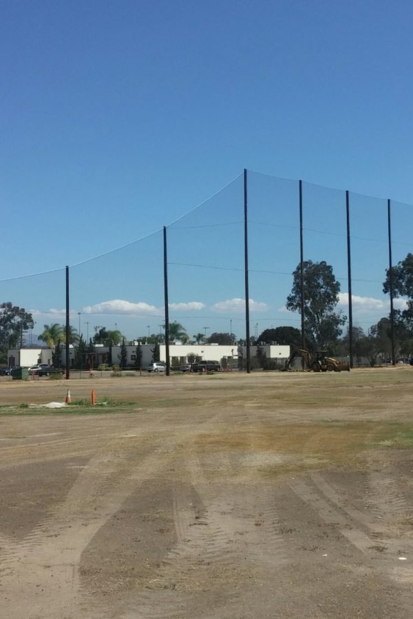 A grassy and dirt field with tall Judge Netting for a sports area in the background. Trees, buildings, and a clear sky are visible, reminiscent of the scenic views at Miramar Memorial Golf Course.