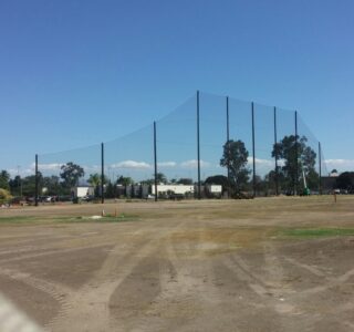 A grassy and dirt field with tall Judge Netting for a sports area in the background. Trees, buildings, and a clear sky are visible, reminiscent of the scenic views at Miramar Memorial Golf Course.