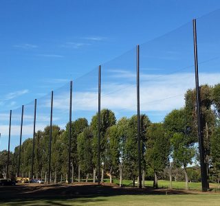 Golf netting poles standing in a line across a grassy park area with trees and a clear blue sky.