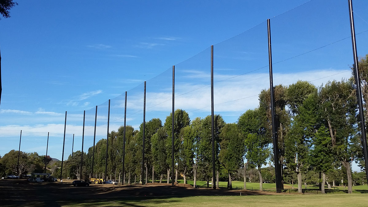 Golf netting poles standing in a line across a grassy park area with trees and a clear blue sky.