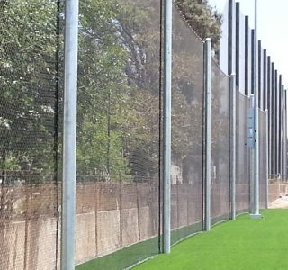 Outdoor cricket training nets alongside a pathway, with high sports netting and lush green grass.