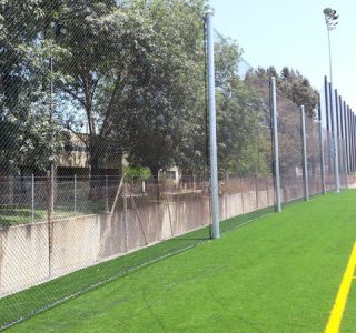 An empty soccer field with artificial turf, marked lines, and surrounded by sports netting, with trees and lampposts in the background.