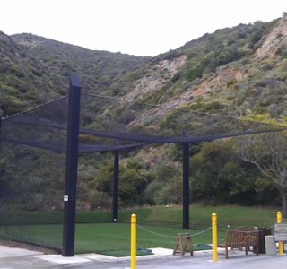 Large empty batting cage surrounded by green mountains in a grassy field, with a bench and industrial netting barricades nearby.