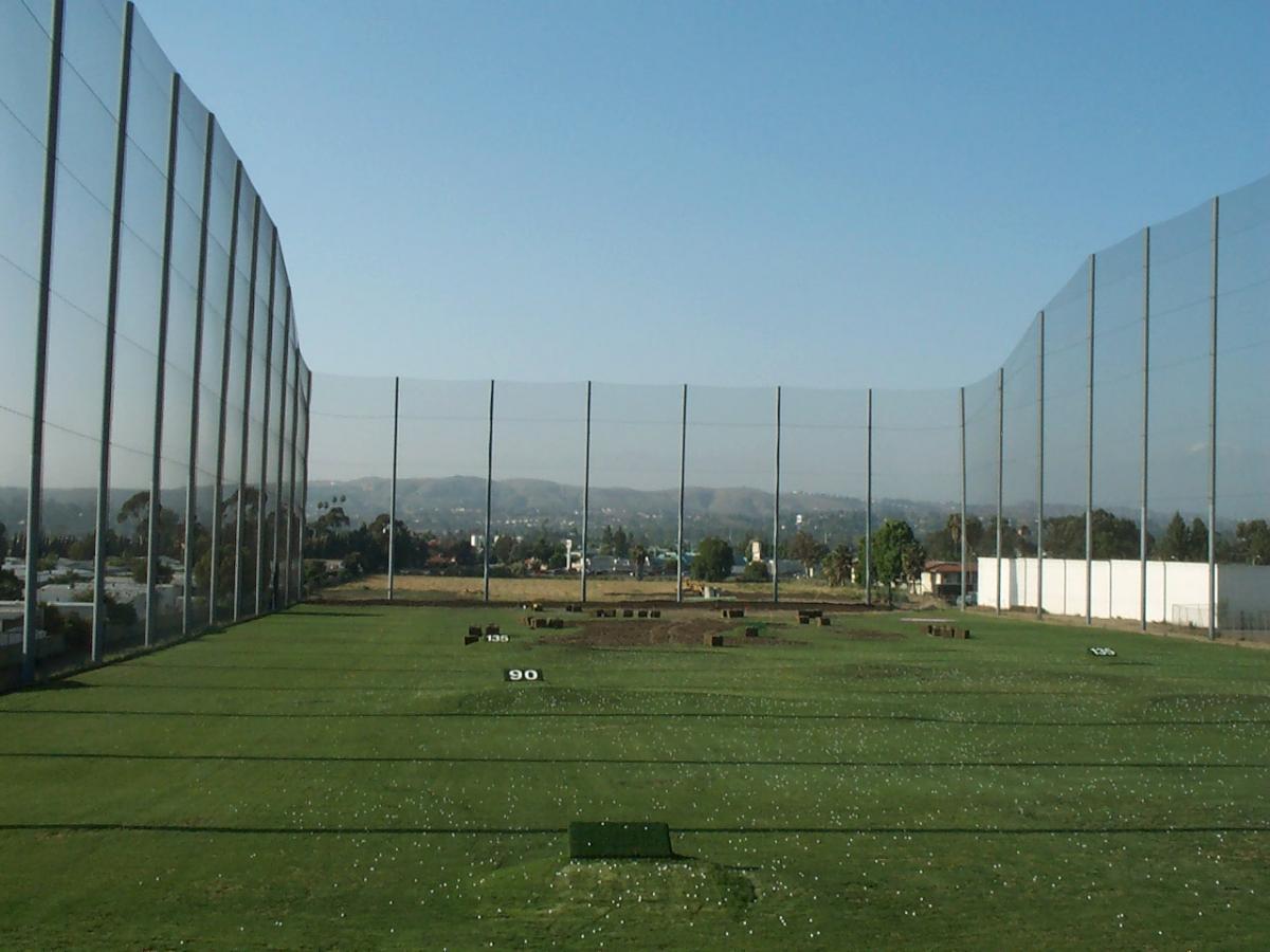 A golf driving range with large industrial netting against a blue sky, with a scenic hilly backdrop reflected on the shiny surface of the nets.