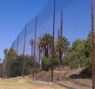 Reflective sound barrier wall beside a road with images of palm trees and a grassy hill mirrored on its surface, enhanced with industrial netting for added durability.