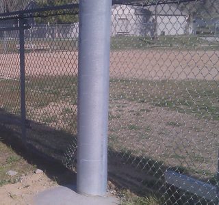 A chain-link fence, commonly used in sports netting, and a metal pole in the foreground with a grassy field and trees in the background.