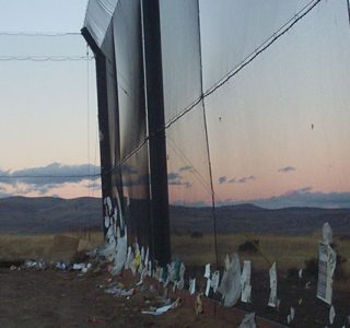 A barbed wire fence with attached scraps of paper and trash, overlooking a vast landscape at dusk, reinforced with industrial netting.