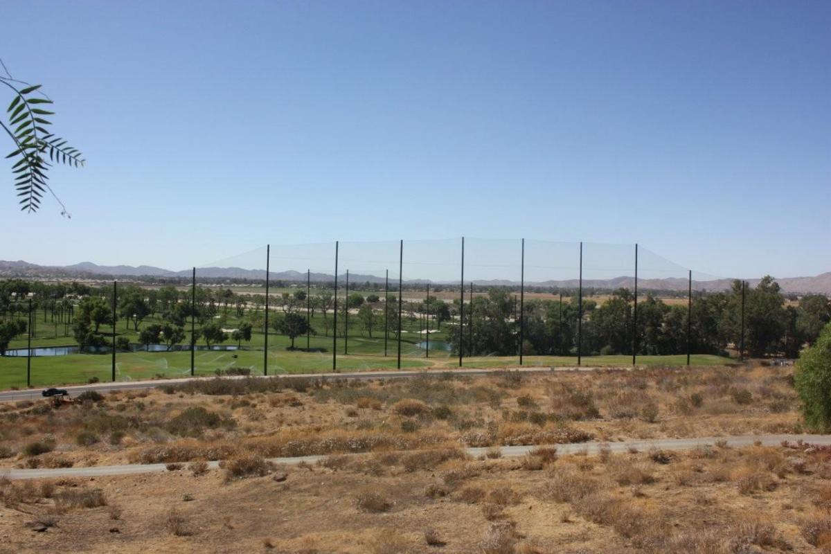 Wide view of a lush golf course with golf netting, surrounded by arid landscape and hills under a clear blue sky.