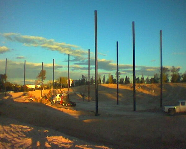 Construction site with tall vertical poles and scattered equipment, set against a backdrop of a sandy terrain and golf netting under a clear sky at dusk.