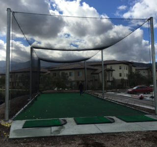 Outdoor batting cage with sports netting and green turf, set against a cloudy sky and residential backdrop, with a person standing nearby.