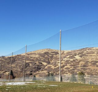 A safety net, designed for golf netting, borders a grassy area with sparse snow patches, overlooking rolling hills under a clear blue sky.