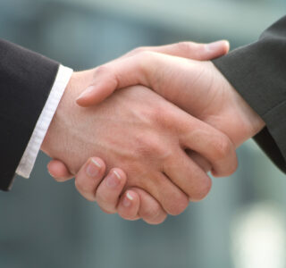 Two people in business attire shaking hands in front of a blurred sports netting background.