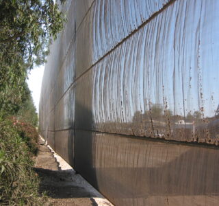 Long view alongside a tall, weathered industrial netting fencing an industrial site, with foliage on the left and clear sky above.