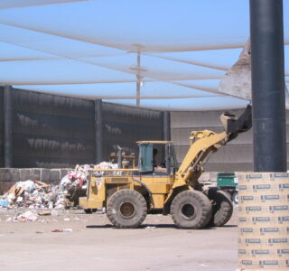 A yellow wheel loader moving trash in a waste management facility, with landfill netting surrounding the area and various debris scattered around under a roof.