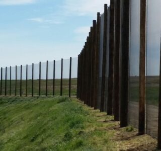 Long metal fence stretching across a grassy landscape under a cloudy sky, bordered by golf netting.