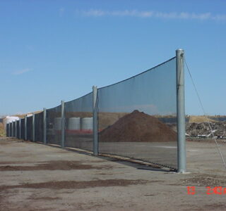 A chain-link fence with several posts and tension wires enclosing a large pile of dirt at an industrial site, covered by landfill netting, with a clear blue sky above.