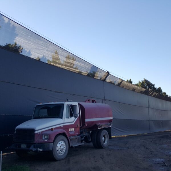 A water truck parked next to a large solar panel installation, secured with industrial netting, on a sloping hillside under a clear sky.