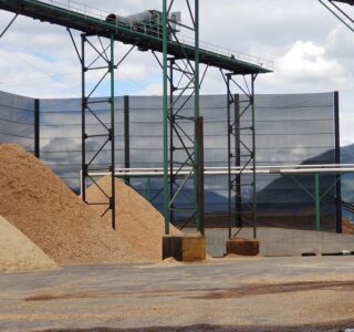 An industrial site with piles of wood chips, metal constructions, and industrial netting covered storage areas under a cloudy sky.