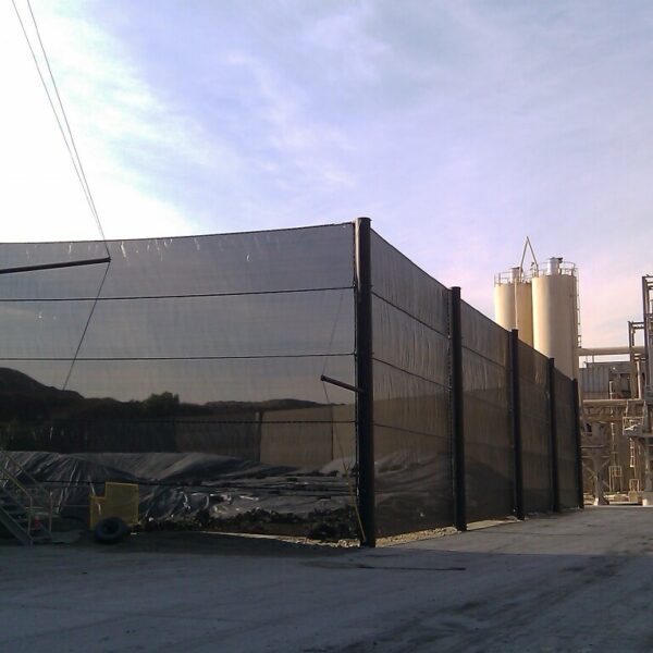 Industrial site with storage silos, industrial netting covering construction materials, and a concrete building under clear skies.