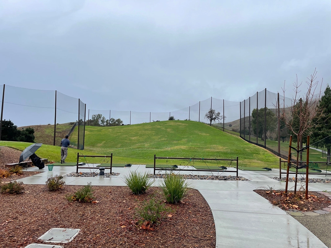 Wet concrete pathway leading to a green grassy hill with a person standing beside tall safety netting at Spring Valley Golf Course on a cloudy day.
