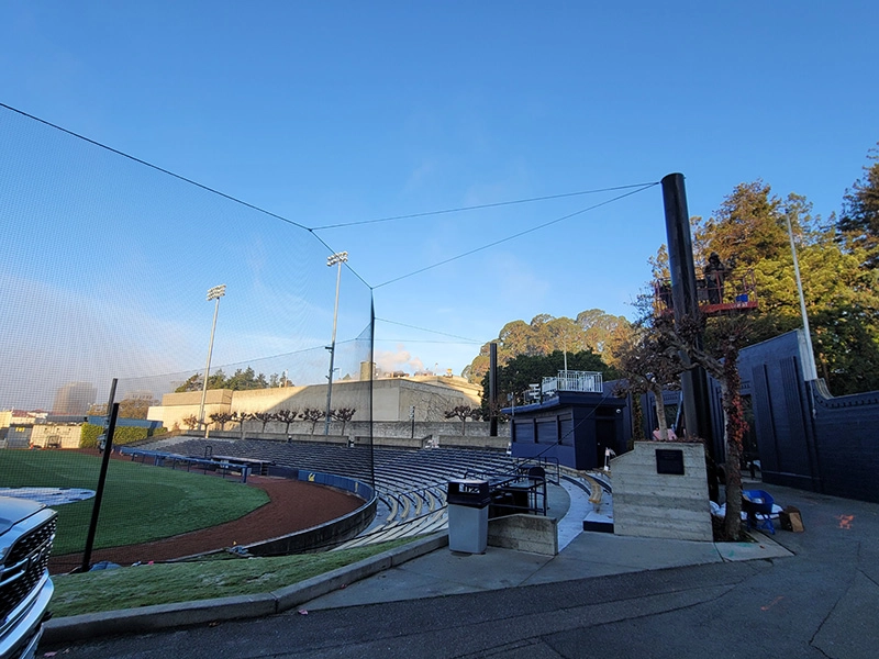 Golden hour sunlight bathes the University of Berkeley baseball field, highlighting the curve of safety netting and the surrounding stadium infrastructure.