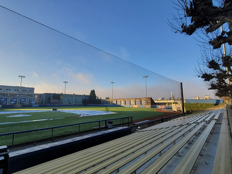 Morning sun illuminates the University of Berkeley Baseball Stadium, showcasing the backstop and protective netting with empty bleachers in the foreground.