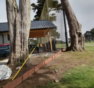 A worker in an orange vest stands near a partially constructed building framed by large trees, reminiscent of Harding Park's scenic surroundings. A wooden beam and netting are set up on a grassy area next to the structure, evoking the meticulous setup of a golf course's first tee.