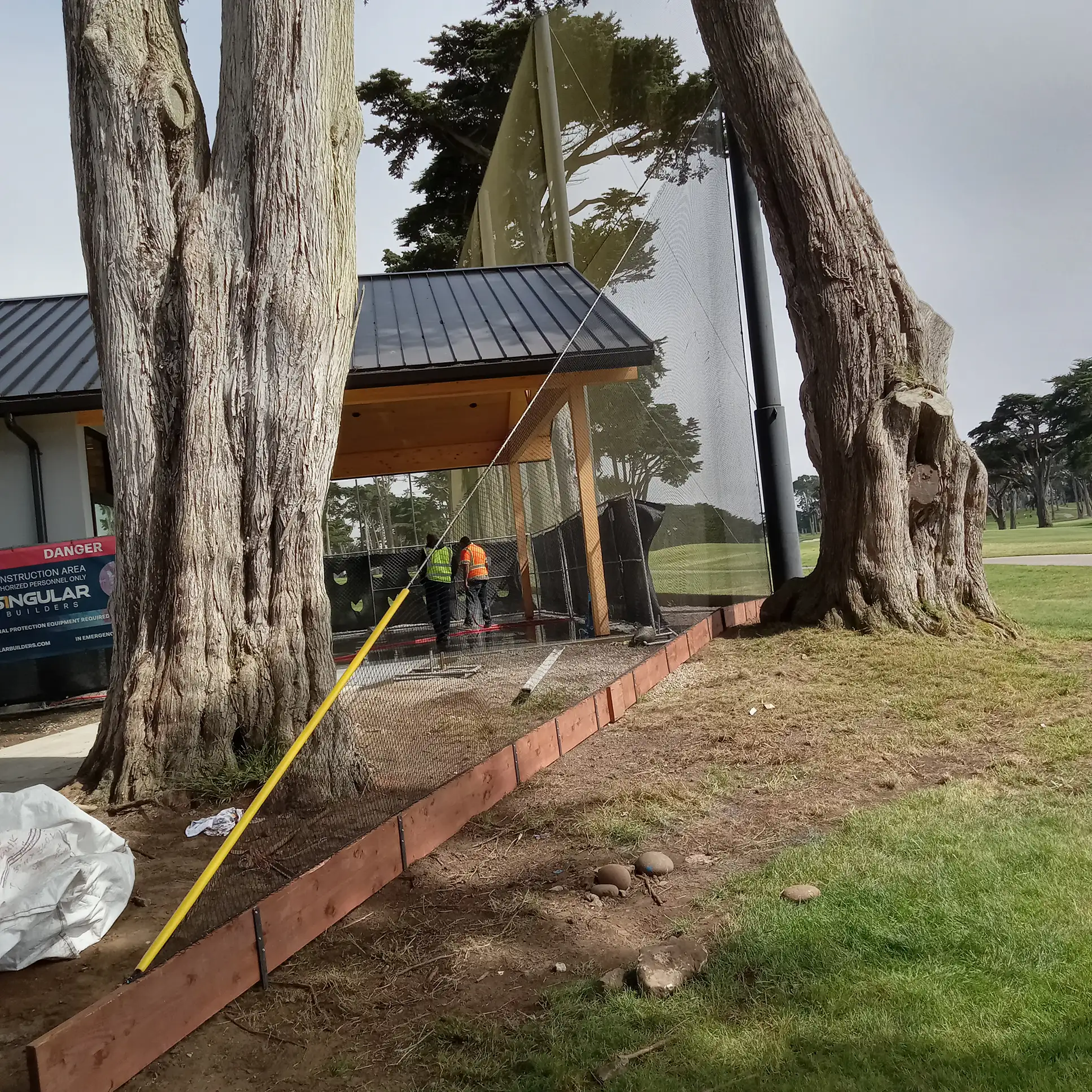 A worker in an orange vest stands near a partially constructed building framed by large trees reminiscent of Harding Parks scenic surroundings A wooden beam and netting are set up on a grassy area next to the structure evoking the meticulous setup of a golf courses first tee