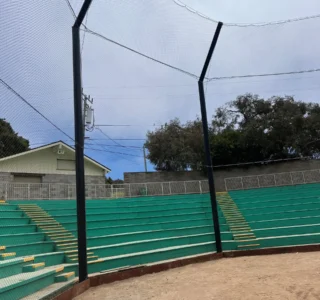 Empty green bleachers and yellow steps are situated behind a protective net at a baseball field, reminiscent of a driving range. The wooden ground is visible in the foreground under a cloudy sky.