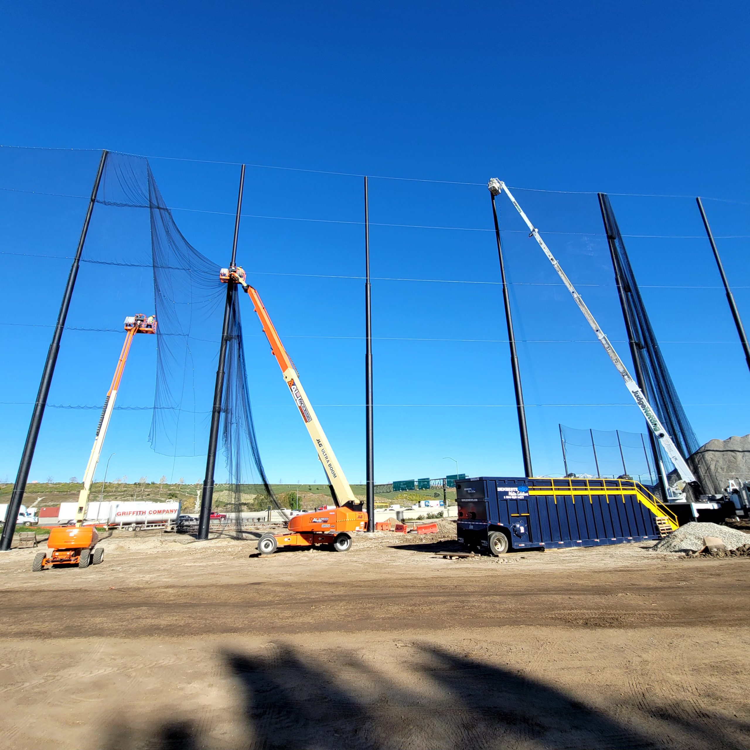 Three cherry pickers are being used to install or repair netting on tall poles at the Diamond Bar Golf Course on a clear blue sky day A large container is placed on the ground near the cherry pickers ensuring the fairway barriers are properly set up