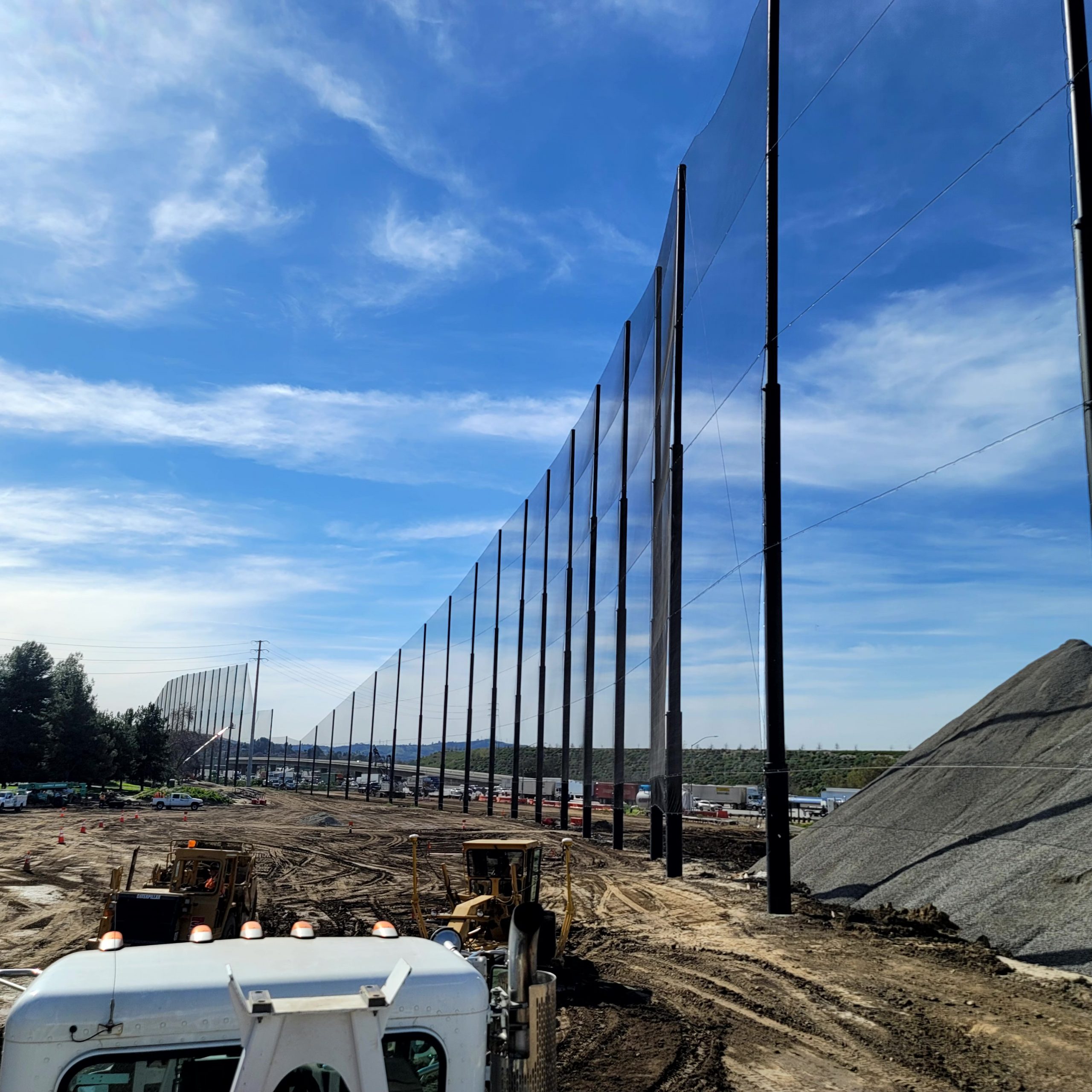 A construction site featuring tall netted poles resembling fairway barriers and machinery beneath a clear blue sky reminiscent of the Diamond Bar Golf Course