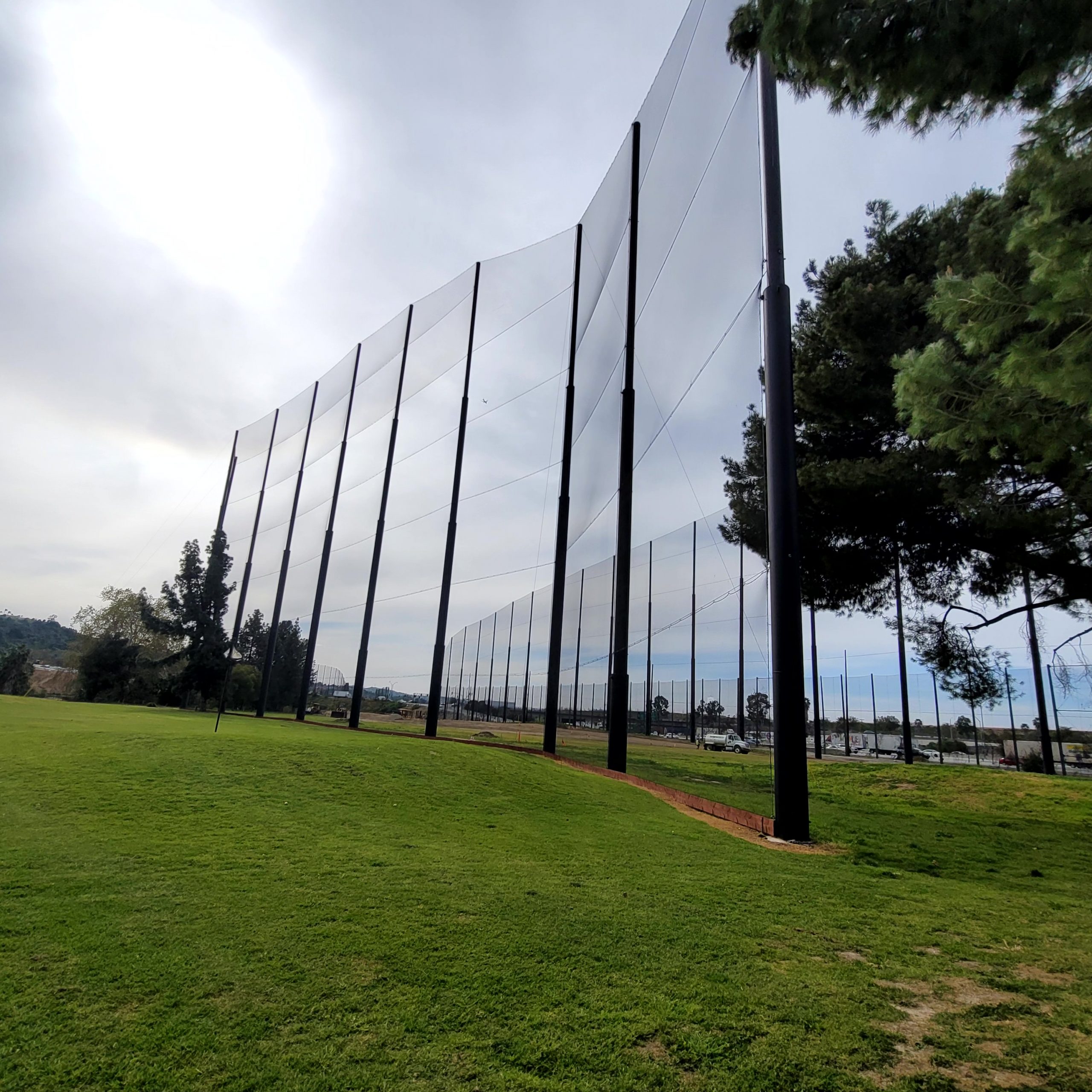 A tall black net fence is installed on a grassy area with trees and an overcast sky in the background serving as one of the Fairway Barriers at Diamond Bar Golf Course