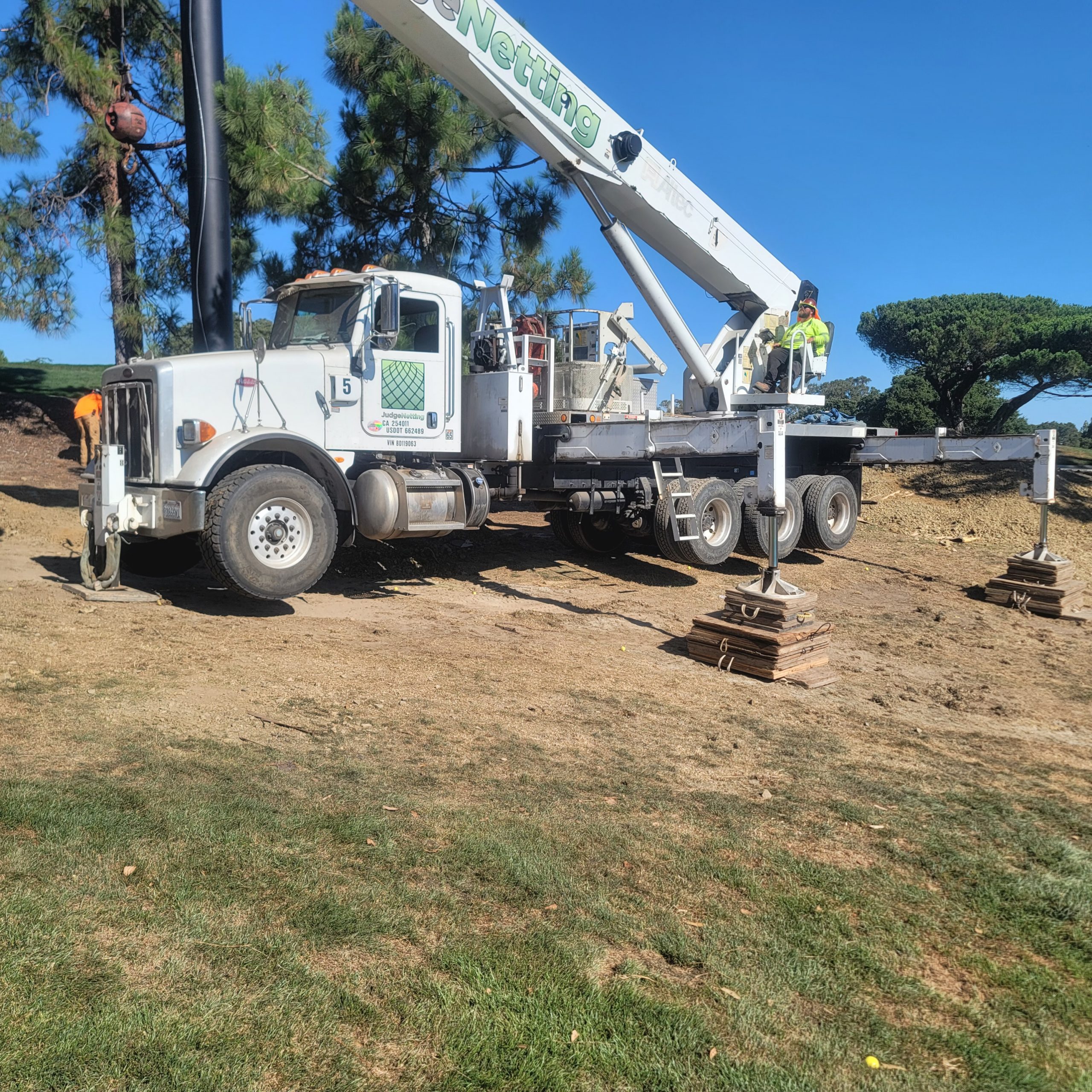 A large truck-mounted crane with extended stabilizers operates near the new range barrier at Peninsula Golf  Country Club with a worker standing by the control panel on a grassy and dirt area