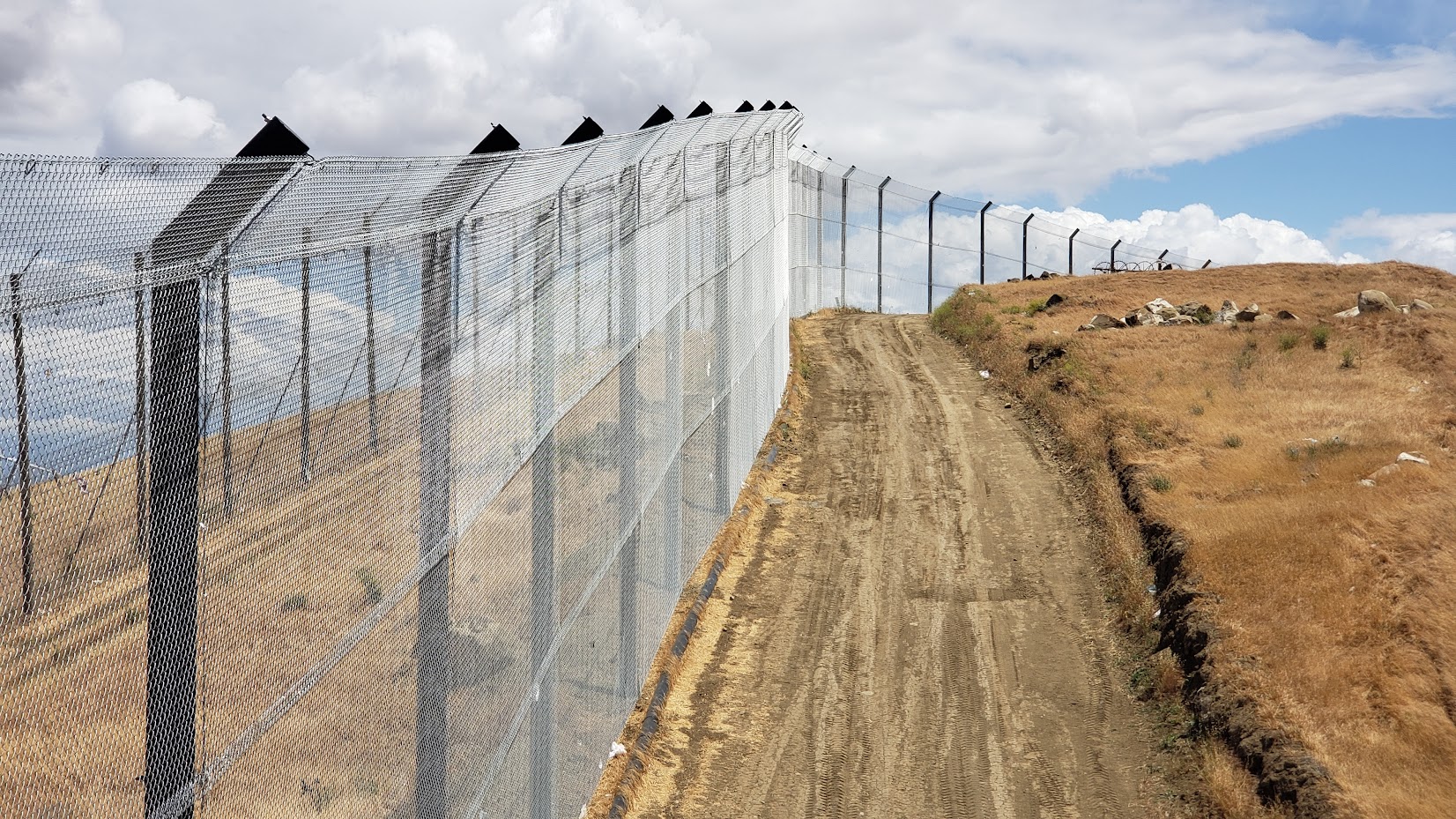 A tall chain-link fence with barbed wire on top runs along a dirt road near Altamont Pass Landfill separating barren land from a cloudy sky backdrop Rocks and dry grass are visible on the right side of the road serving as natural barriers to litter containment