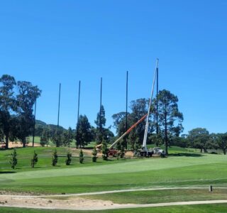 A construction crew using a crane to install tall poles for the New Range Barrier on a grassy field at Peninsula Golf & Country Club, with trees in the background under a clear blue sky.