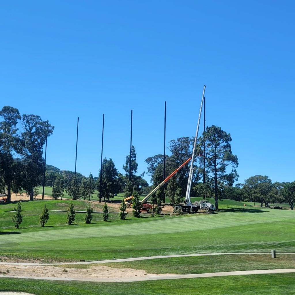 A construction crew using a crane to install tall poles for the New Range Barrier on a grassy field at Peninsula Golf  Country Club with trees in the background under a clear blue sky