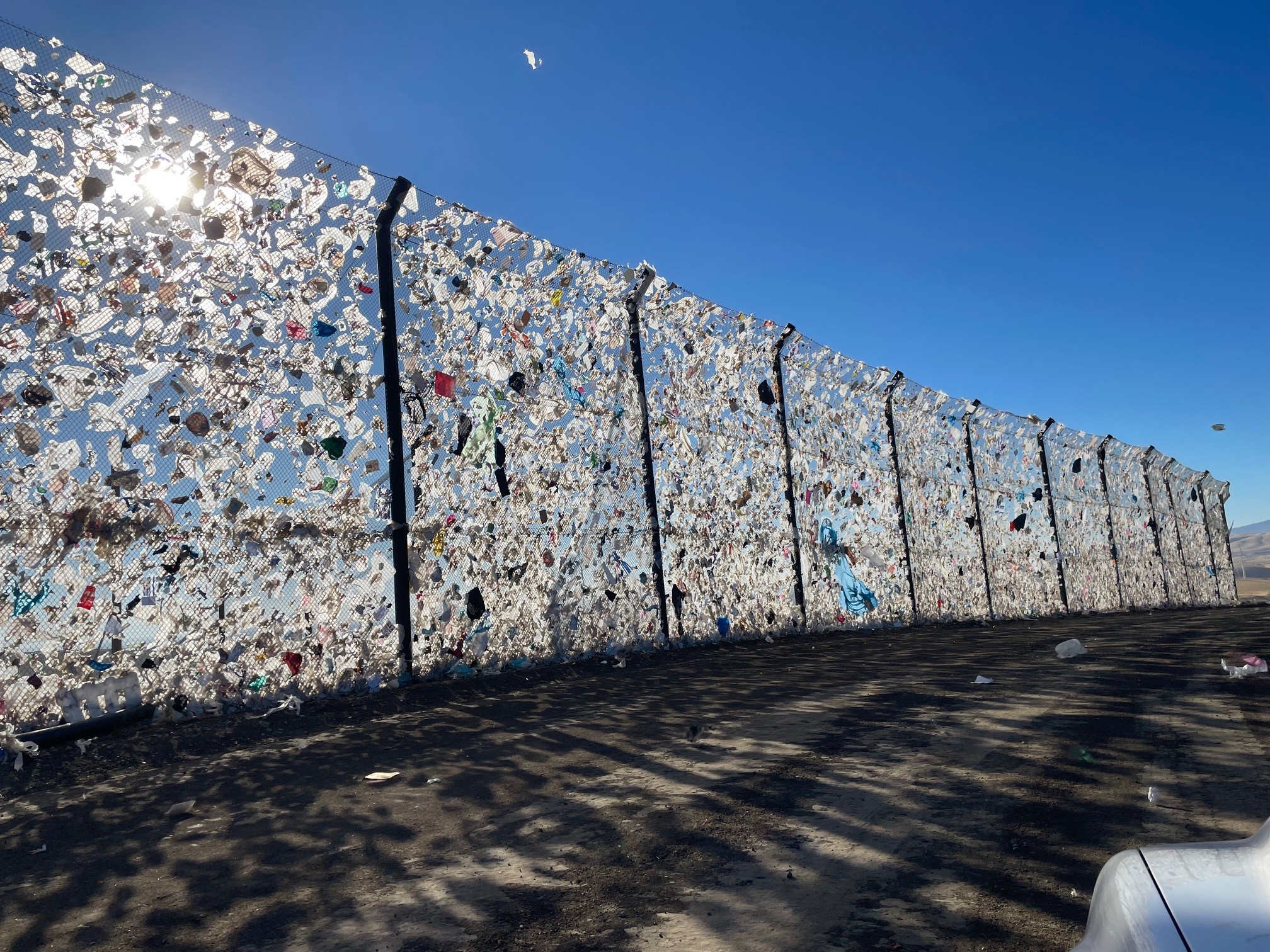 A chain-link fence covered with plastic debris and litter set against a clear blue sky with shadows cast on the ground serves as one of the litter containment barriers near Altamont Pass Landfill