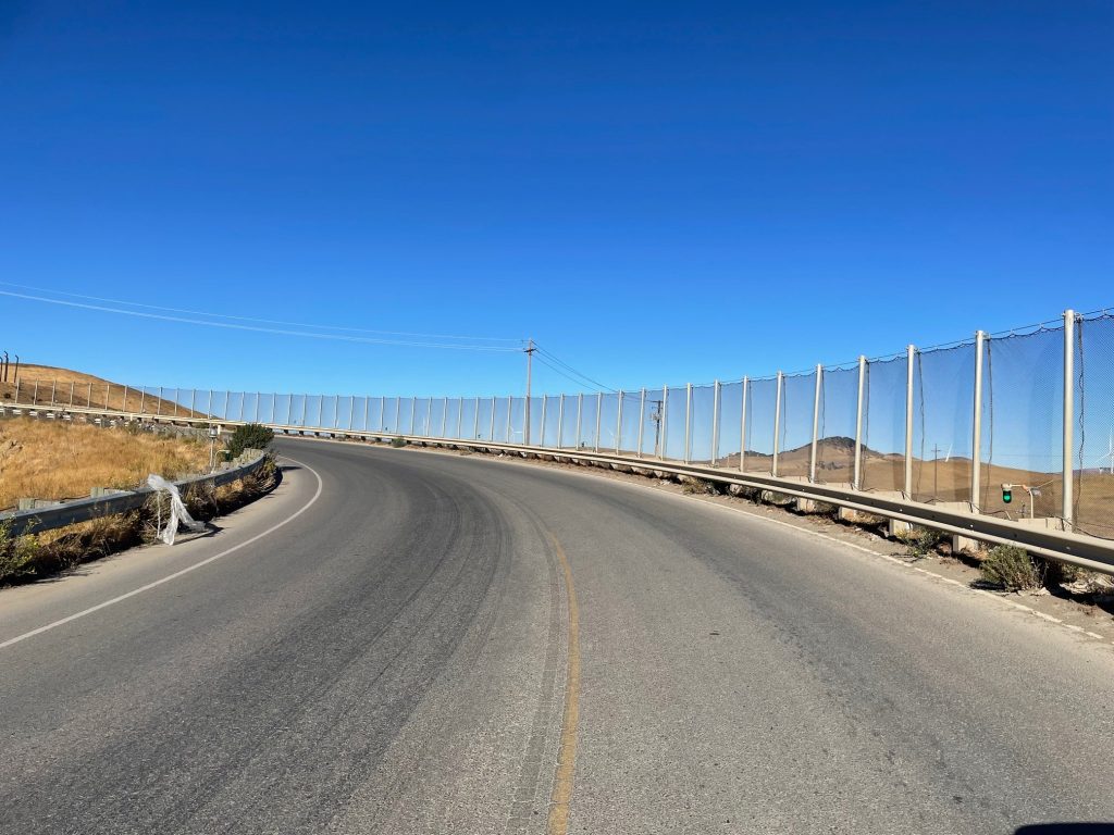 A curved road with guardrails and fencing on the right side, leading into a hilly, arid landscape under a clear blue sky, meanders near the Altamont Pass Landfill where barriers ensure effective litter containment.