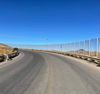 A curved road with guardrails and fencing on the right side, leading into a hilly, arid landscape under a clear blue sky, meanders near the Altamont Pass Landfill where barriers ensure effective litter containment.