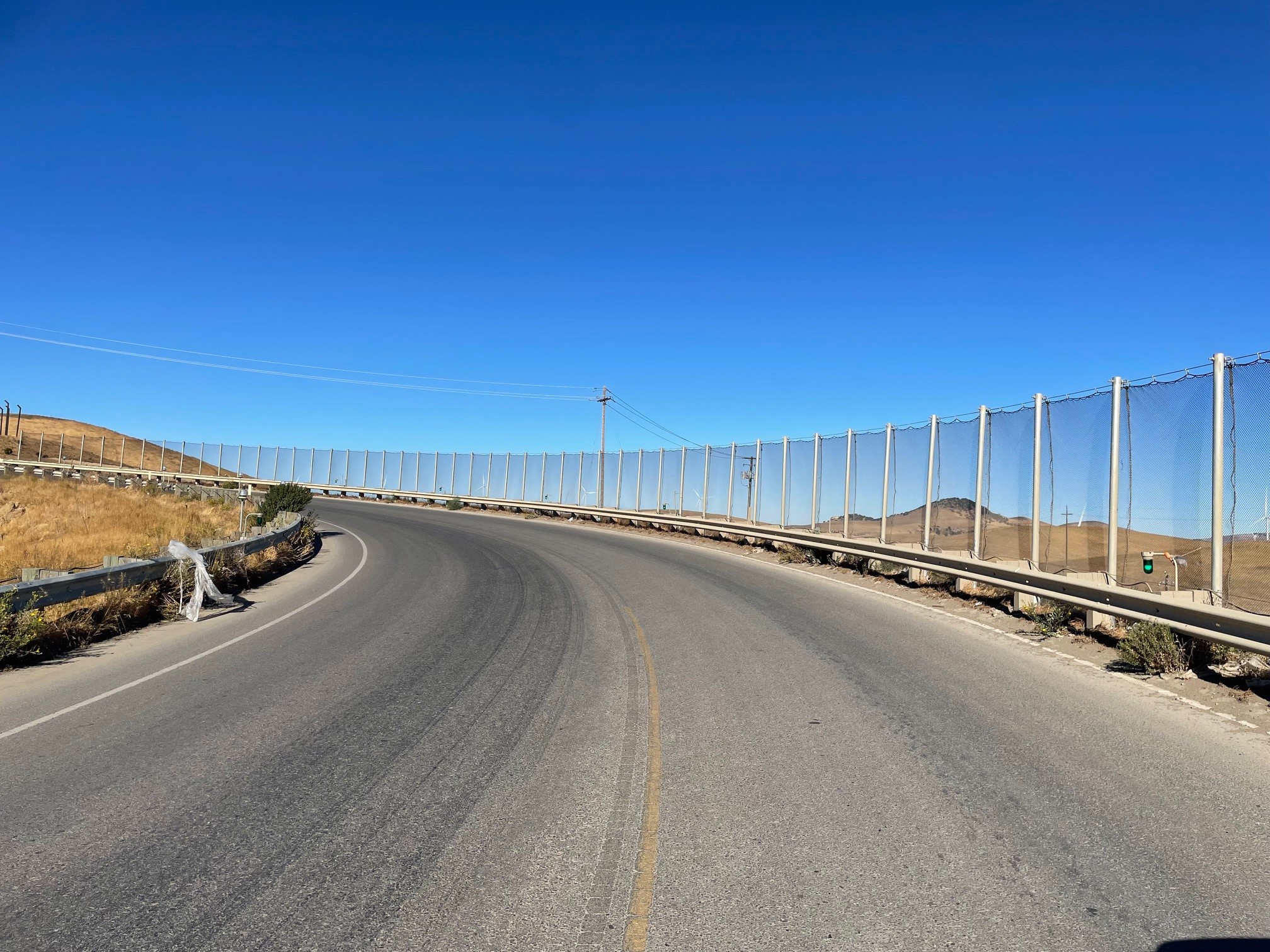 A curved road with guardrails and fencing on the right side leading into a hilly arid landscape under a clear blue sky meanders near the Altamont Pass Landfill where barriers ensure effective litter containment