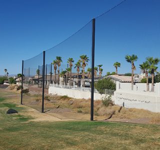A sunlit golf course with palm trees, surrounded by high golf netting, and a residential area in the background.