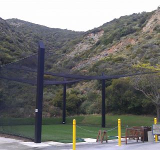 An outdoor batting cage with industrial netting setup in a mountainous park area, featuring nearby benches and yellow barricades.