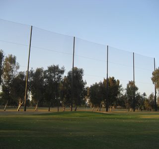 A driving range with tall sports netting, trees in the background, and grass-covered ground under evening light.