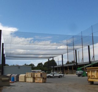 Outdoor industrial area with sports netting overhead, dumpsters, a white truck, and a cloudy sky.