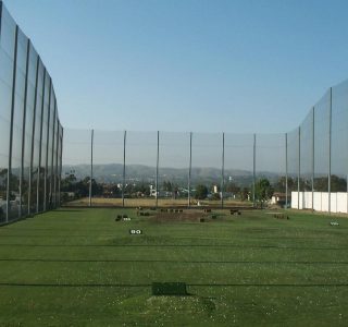 A golf driving range with large industrial netting against a blue sky, with a scenic hilly backdrop reflected on the shiny surface of the nets.