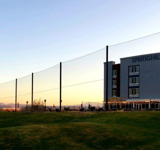 Sunset view of Springhill Suites hotel with industrial netting and people on a grassy area, with distant mountains under a clear sky.