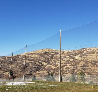 A safety net, designed for golf netting, borders a grassy area with sparse snow patches, overlooking rolling hills under a clear blue sky.