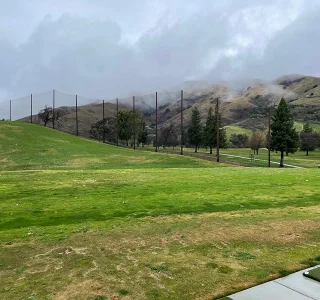 Overcast sky above the vibrant green fairway of Spring Valley Golf Course, with tall safety netting posts lined up along the slope.