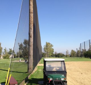 Golf driving range with a utility cart parked beside a tall sports netting, under a clear blue sky.