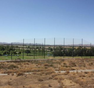 Wide view of a lush golf course with golf netting, surrounded by arid landscape and hills under a clear blue sky.